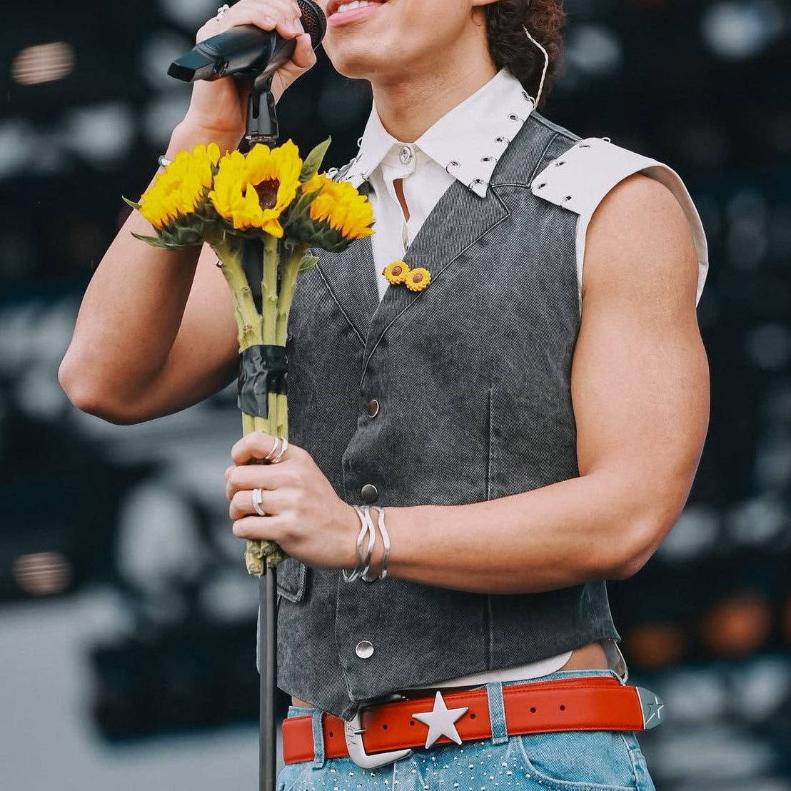 Person with a red leather belt holding a microphone and flowers with a blurred background