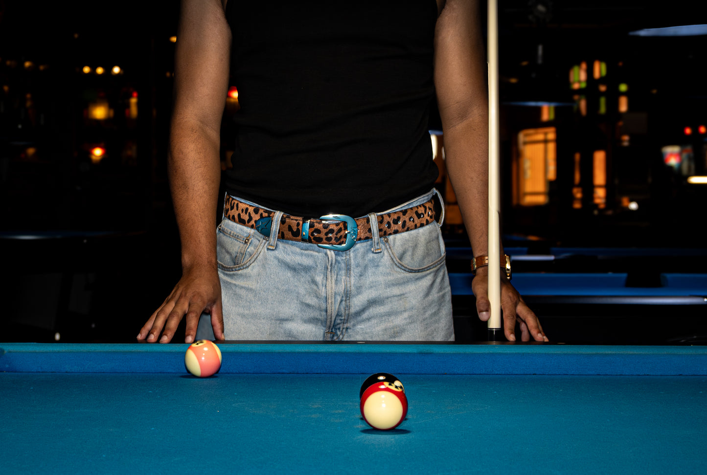 A person with a leopard-printed belt, standing behind a pool table with two balls on the surface, blurred city lights in the background.