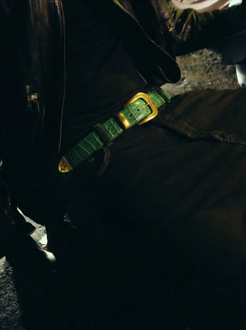Close-up of a green crocodile-printed belt with a gold buckle on dark clothing.