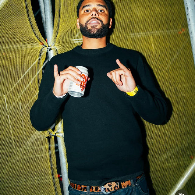 A man in a black sweater with a leopard print belt, holding a cup with a yellow background