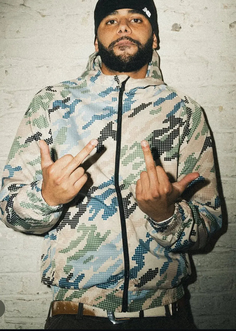 Person wearing a camouflage jacket wiith a cowhide leather belt making hand gestures against a textured wall.