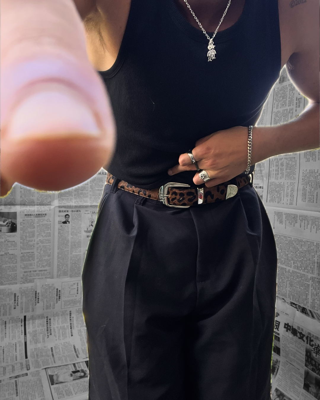 Person wearing a black tank top and dark pants with a leopard print belt, standing against a newspaper background.