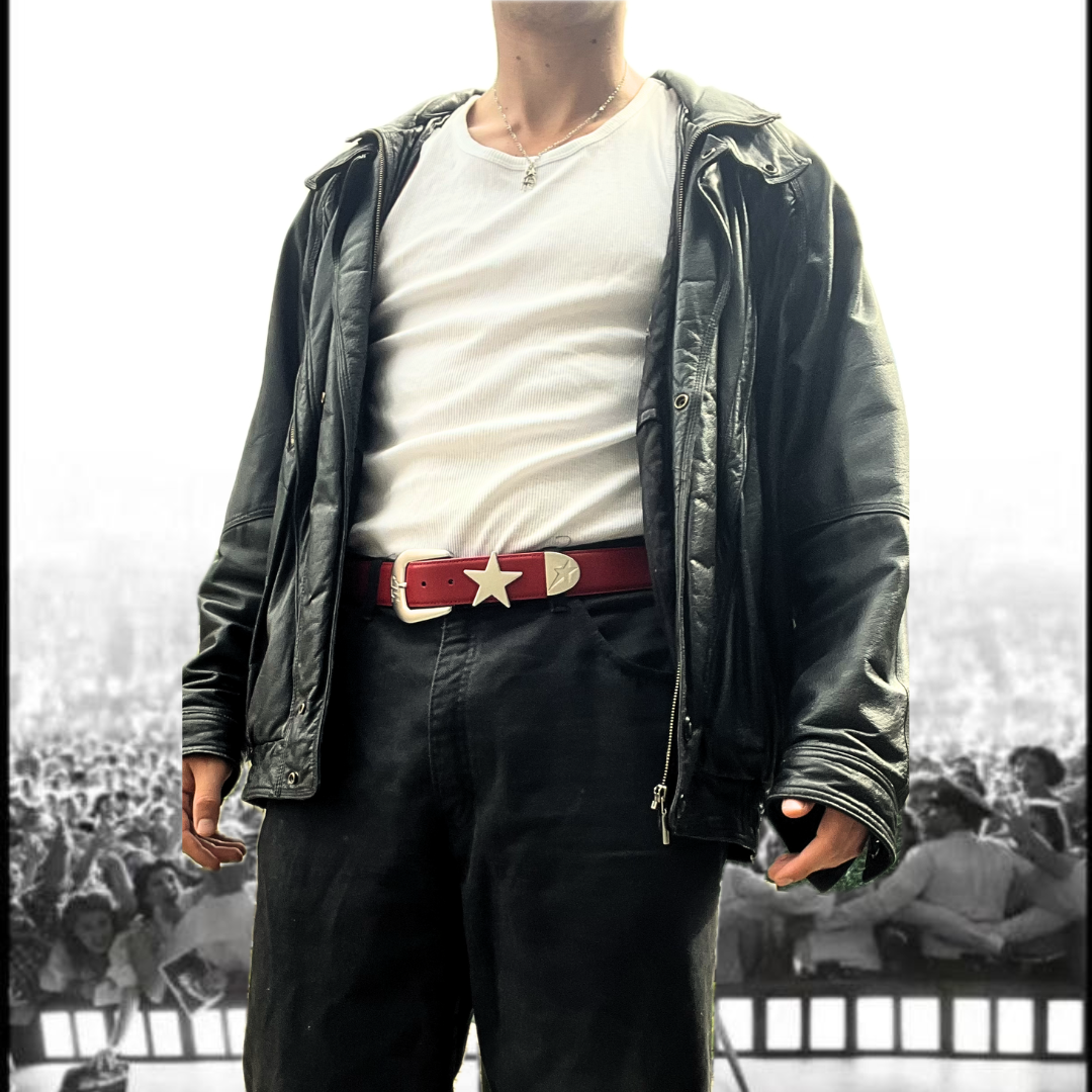 Person wearing a black leather jacket, white shirt, and dark pants with a red belt in a blurred indoor setting.