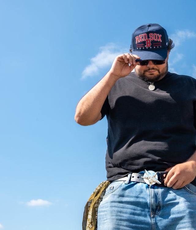 A man wearing a 'Red Sox' cap and a spinnable star leather belt against a blue sky
