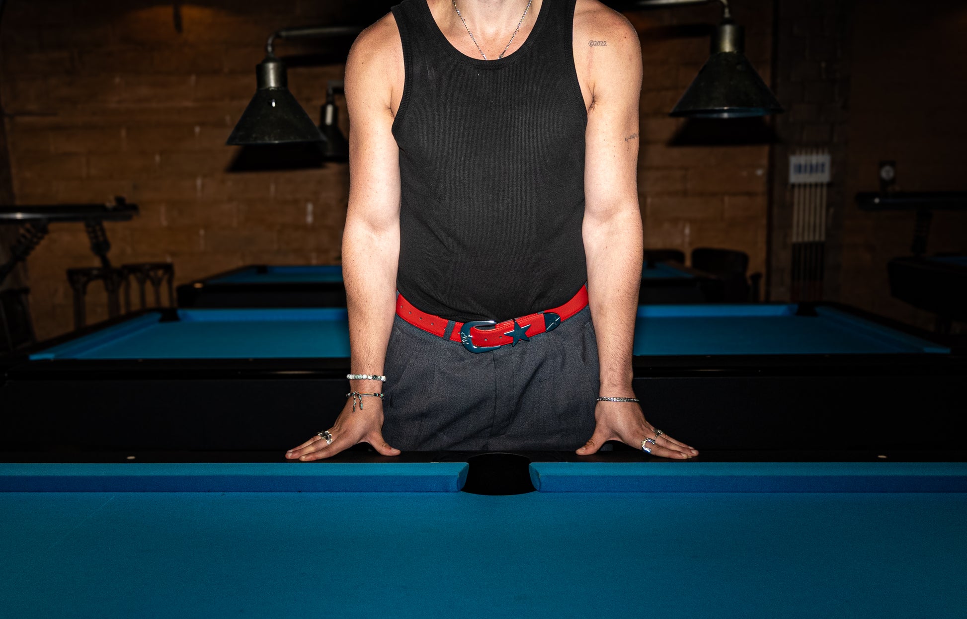 Person standing on a pool table wearing a black tank top and gray shorts with a red belt.